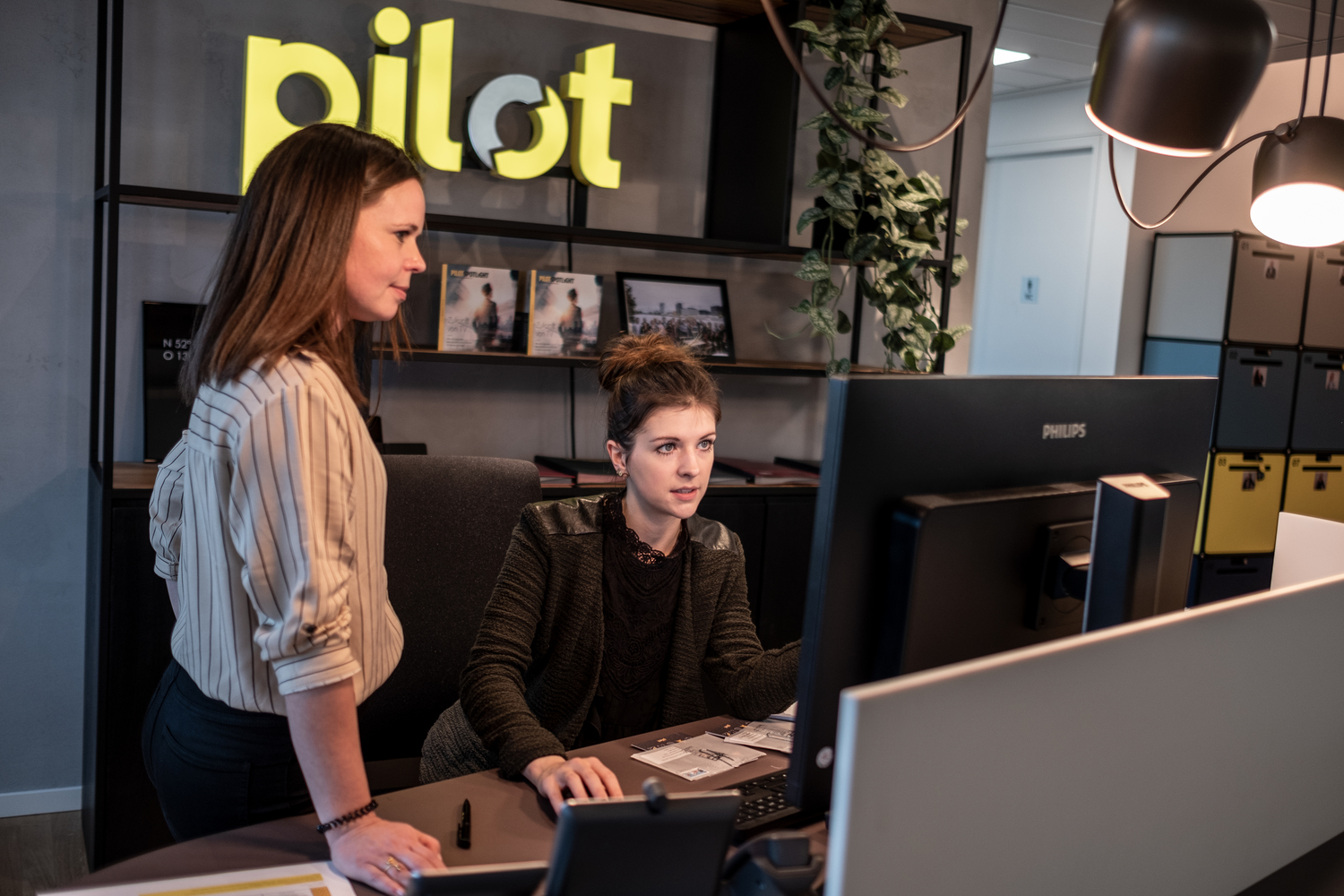 Zwei Personen arbeiten an einem Computer mit 'pilot'-Logo im Hintergrund.