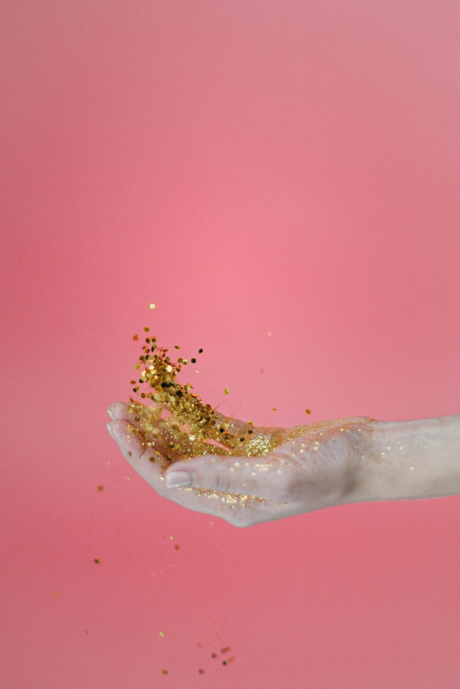 Eine Hand hält goldenen Glitzer vor einem rosa Hintergrund.