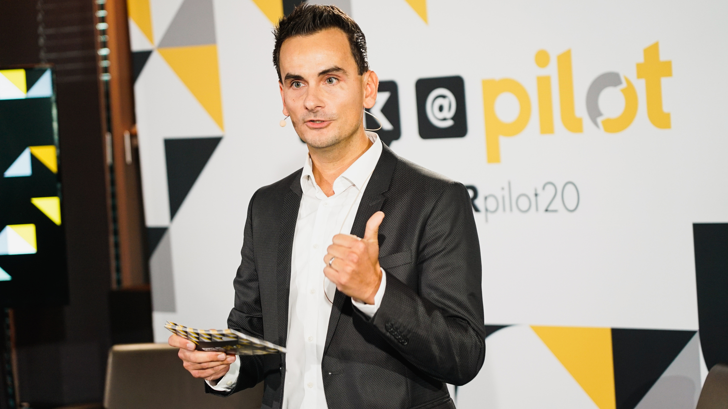 Ein Mann in einem dunklen Anzug hält Karten in der Hand und spricht auf einer Bühne mit dem „pilot“ Logo im Hintergrund.