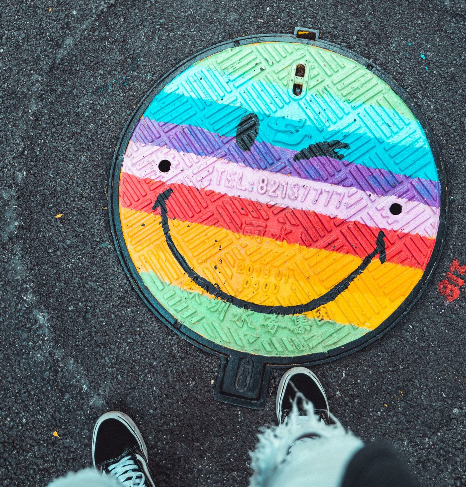 Ein bemalter Kanaldeckel mit einem Regenbogen-Smiley.
