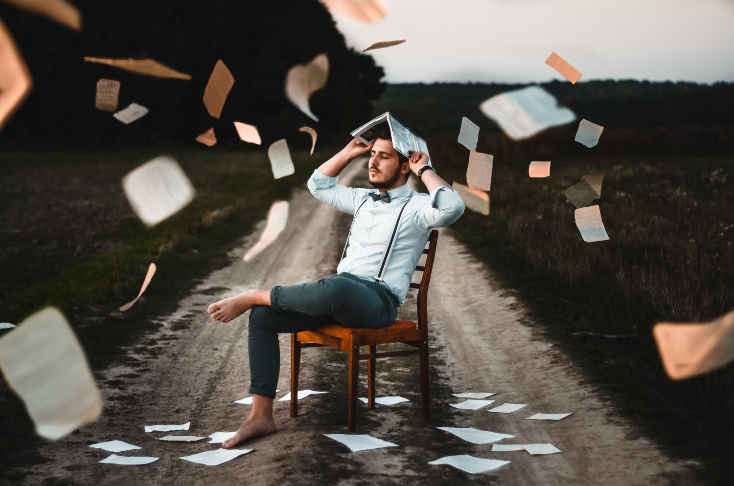 Dmitry Ratushny O33IVNPb0RI Unsplash 1 Scaled 1 Ein Mann sitzt auf einem Stuhl mit einem Buch auf dem Kopf, umgeben von fliegenden Blättern.
