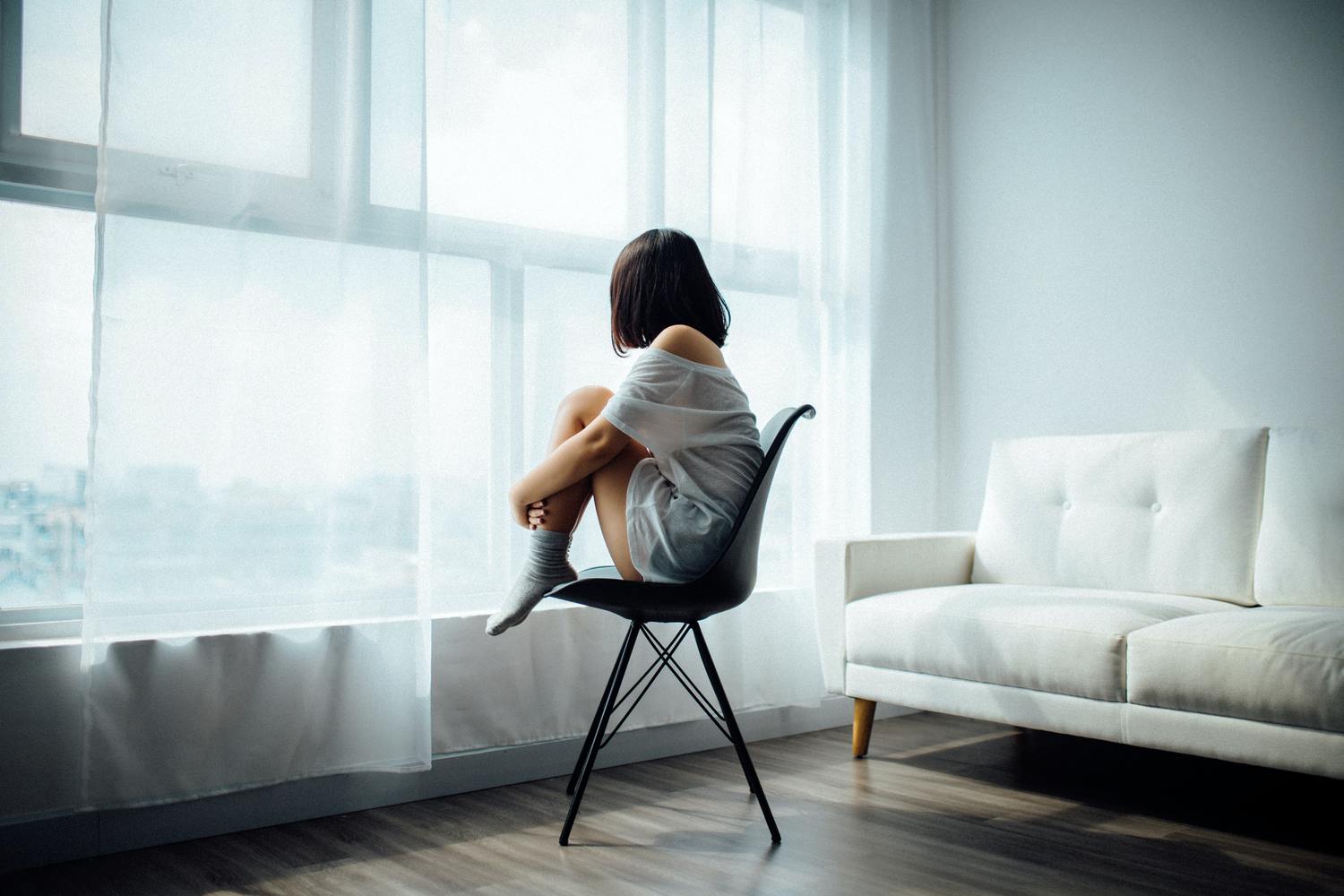 Anthony Tran VXymirxr5ac Unsplash Scaled 1 Eine Frau sitzt nachdenklich auf einem Stuhl und blickt aus dem Fenster in einem minimalistischen, hellen Raum.