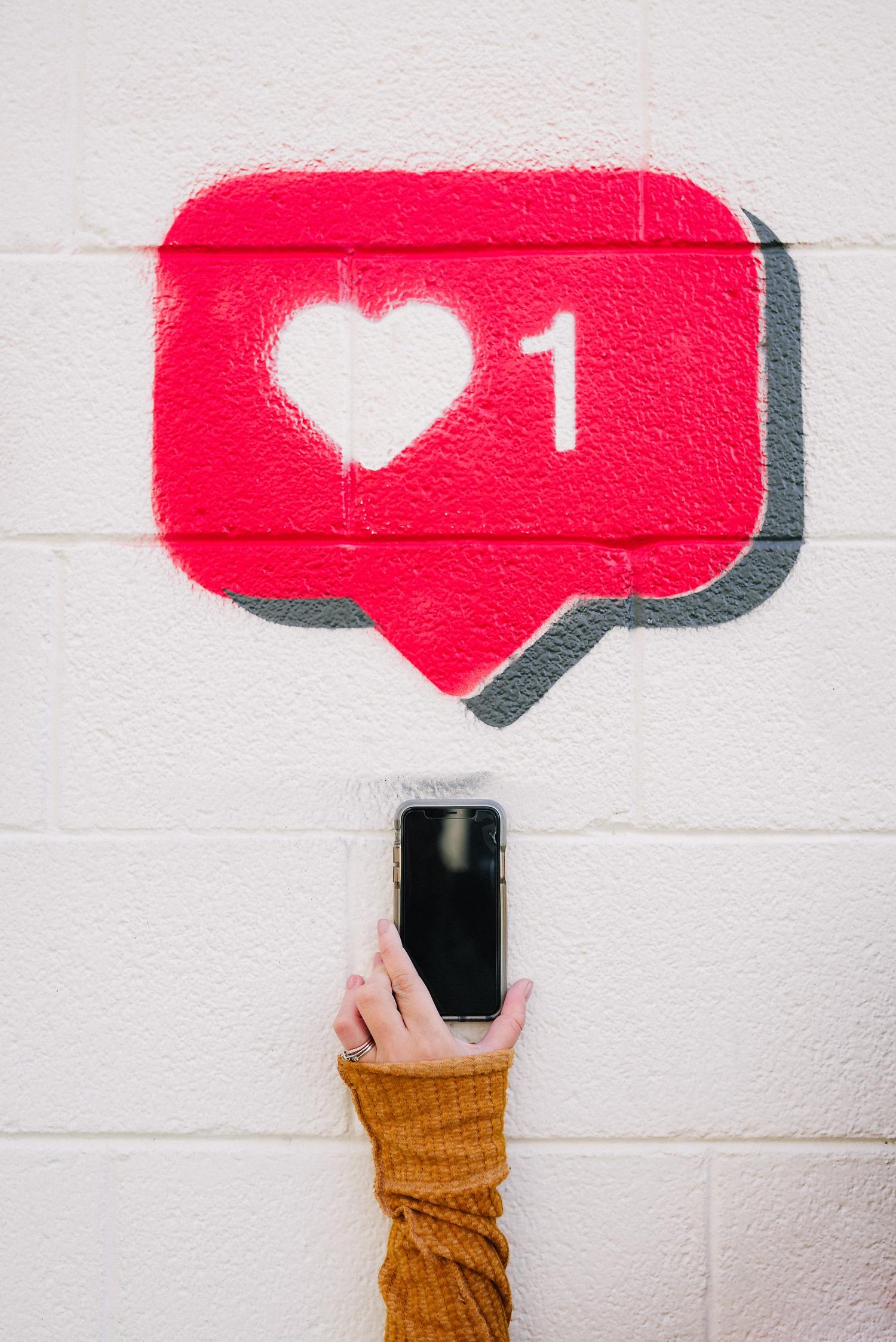 Eine Hand hält ein Smartphone vor eine weiße Wand mit einem aufgesprühten roten Like-Symbol, das ein Herz und die Zahl 1 zeigt.