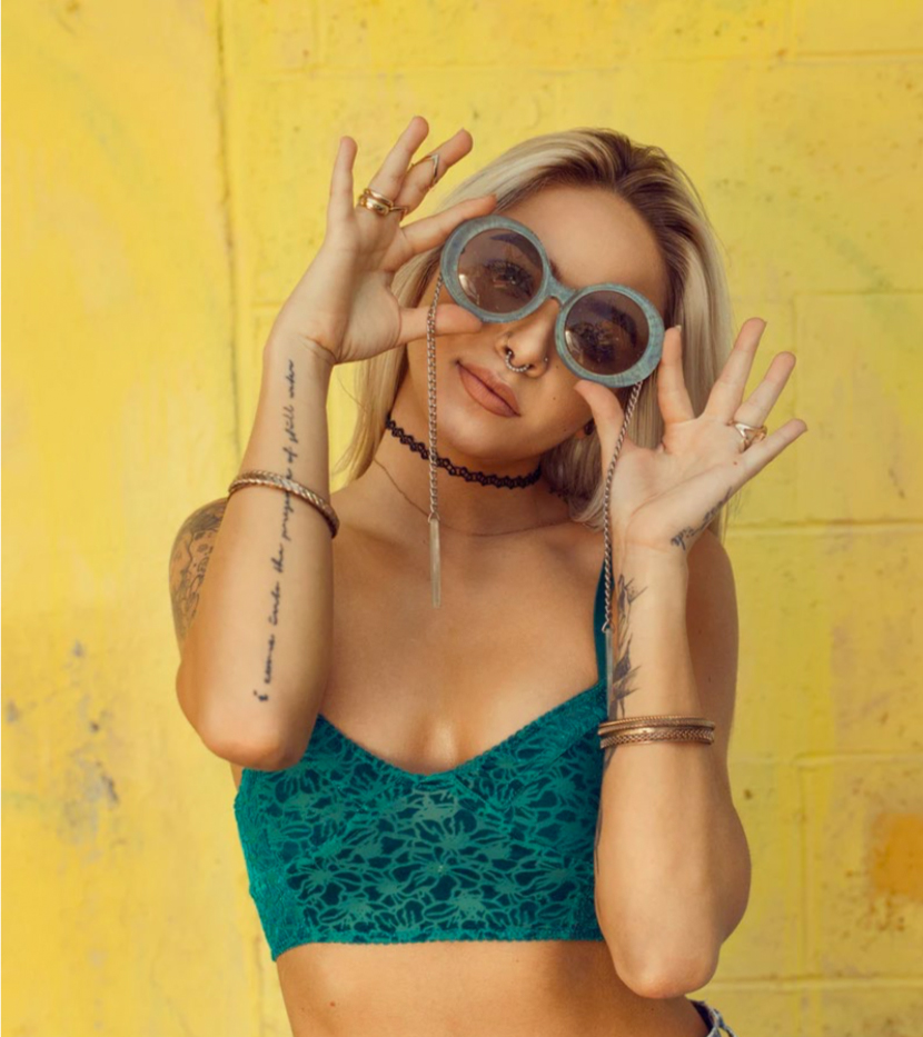 Frau posiert vor einer gelben Wand mit Sonnenbrille.