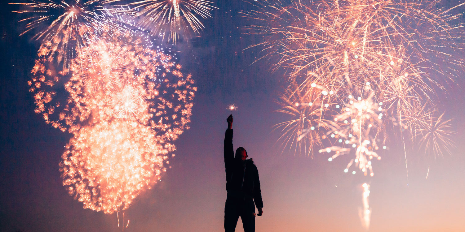 Person hält eine Wunderkerze vor einem Feuerwerk.