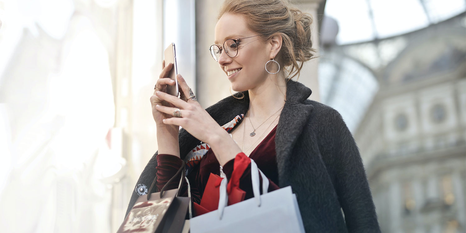2021 MediaTrends RetailRevolution Titel 1 Frau mit Einkaufstaschen schaut auf ihr Smartphone.