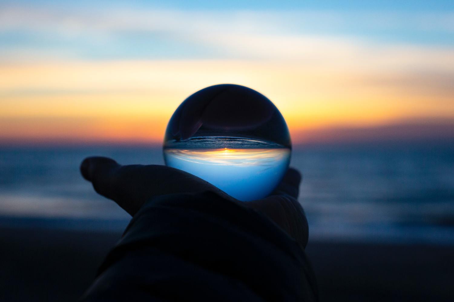 Drew Beamer XU5Mqq0Chck Unsplash Scaled 1 Hand hält eine Glaskugel mit dem Sonnenaufgang im Hintergrund.
