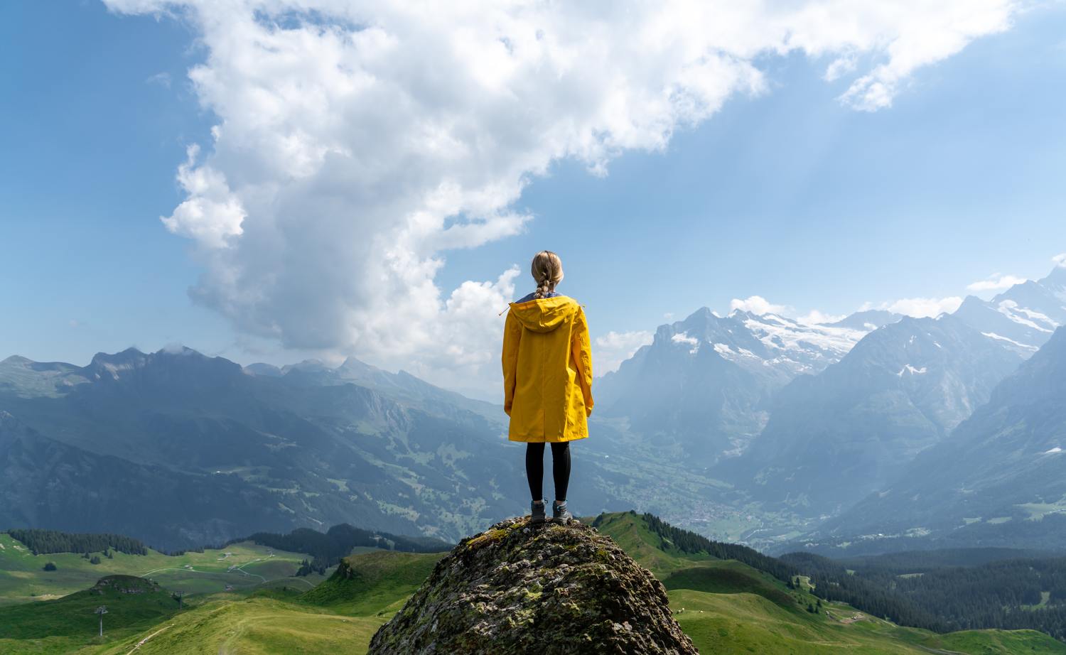 Person in einem gelben Regenmantel steht auf einem Felsen mit Bergen im Hintergrund.