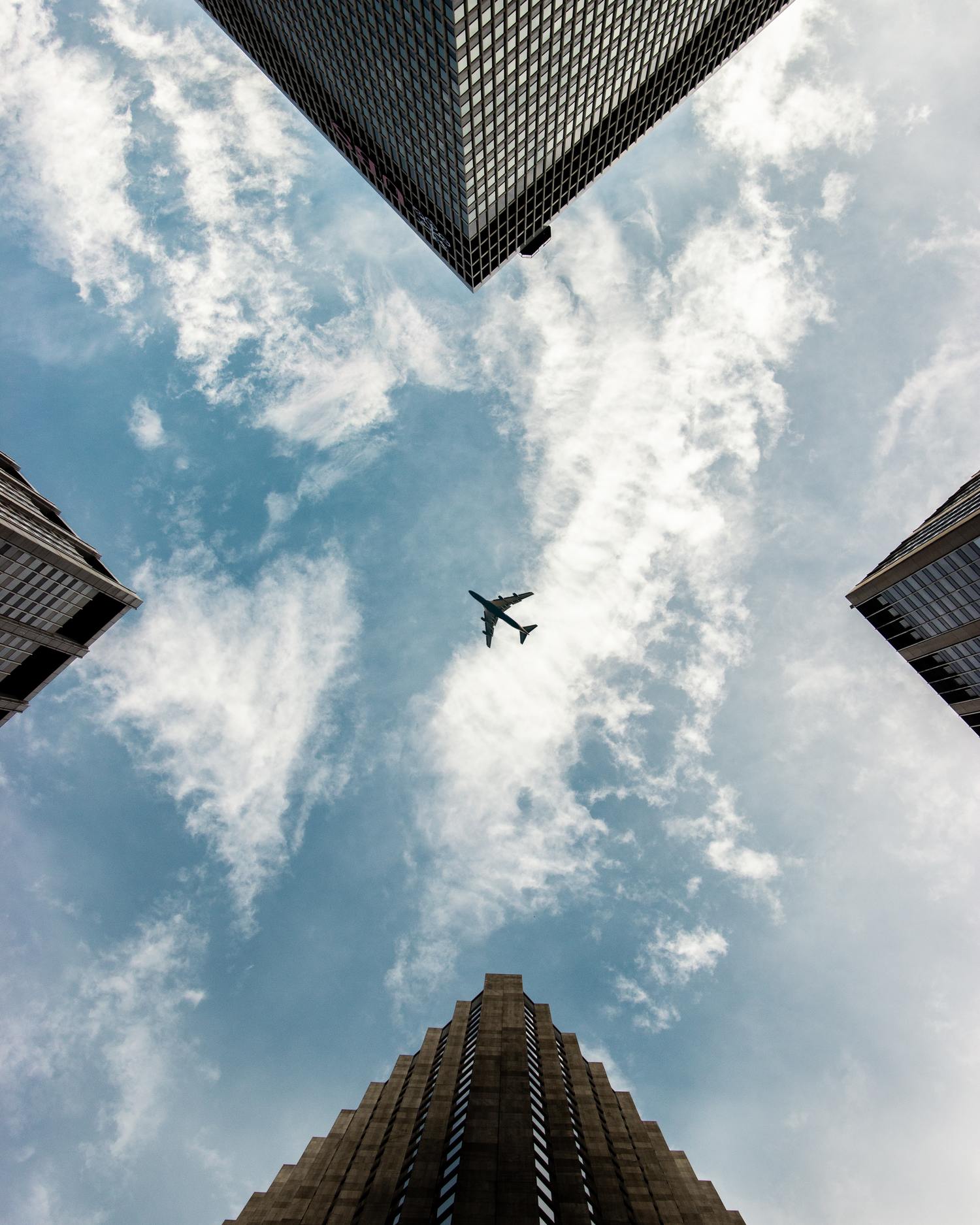 Flugzeug fliegt über Wolken, von hohen Gebäuden umrahmt.