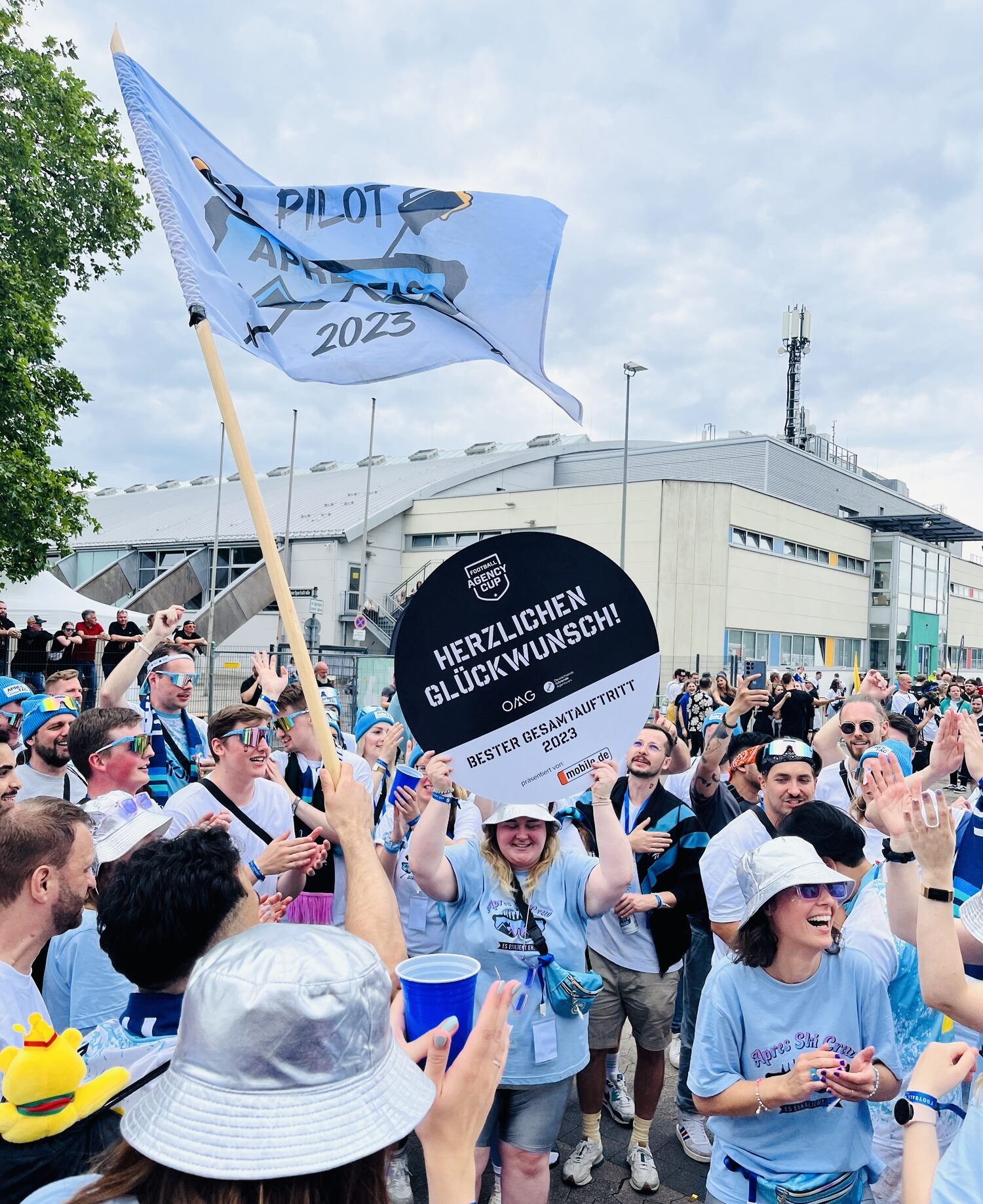 Eine Gruppe von Menschen präsentiert stolz eine blau-weiße Flagge auf dem Agency Cup