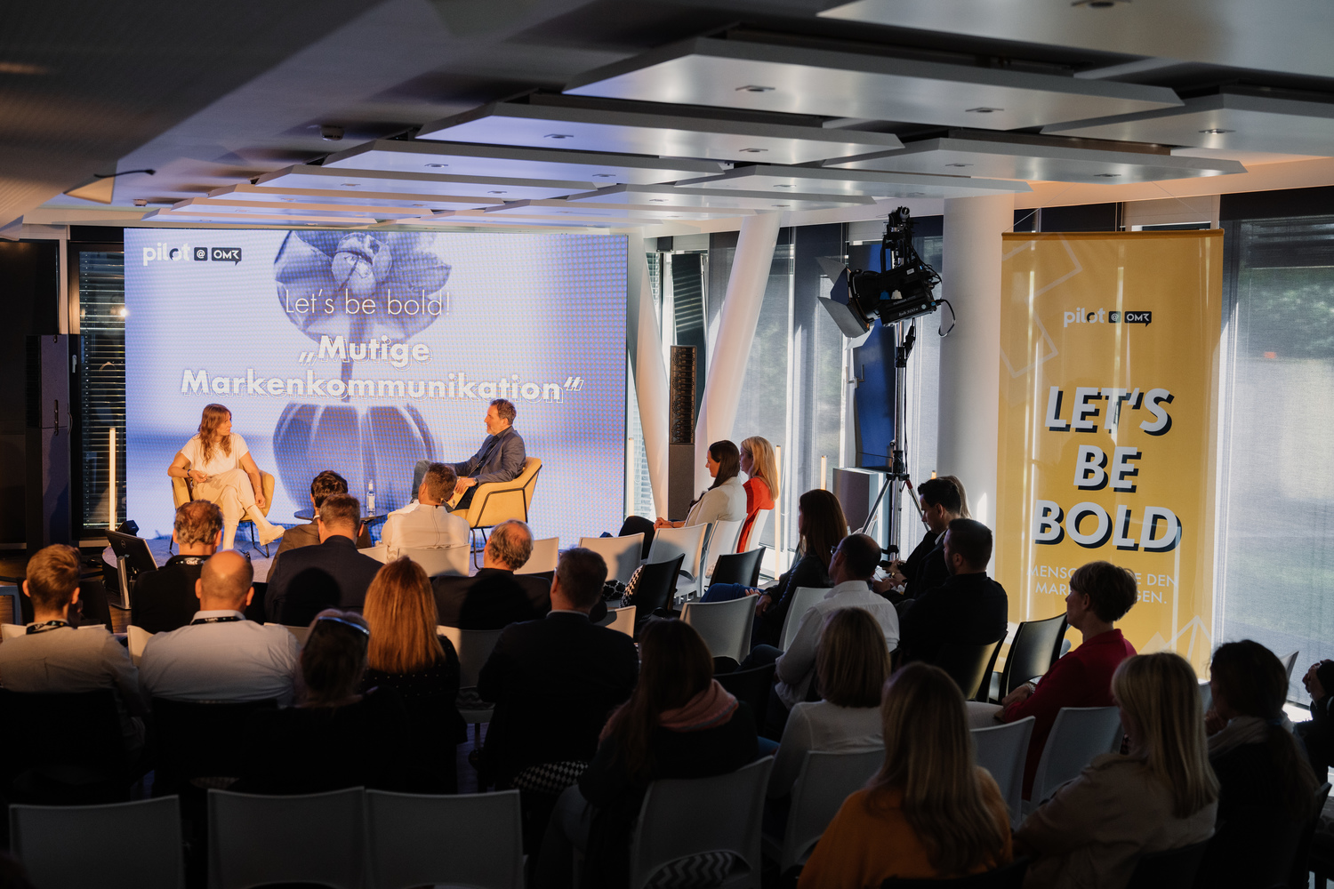 Eine Gruppe von Personen sitzt in Stühlen vor einem Bildschirm und verfolgt aufmerksam die Diskussion auf dem OMR Event bei pilot.
