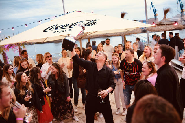 Menschen stehen auf der Tanzfläche im Beachclub bei der Sommerparty 2024. Eine Person steht in der Mitte und streckt den Pokal des Football Agency Cup 2024 in die Luft.