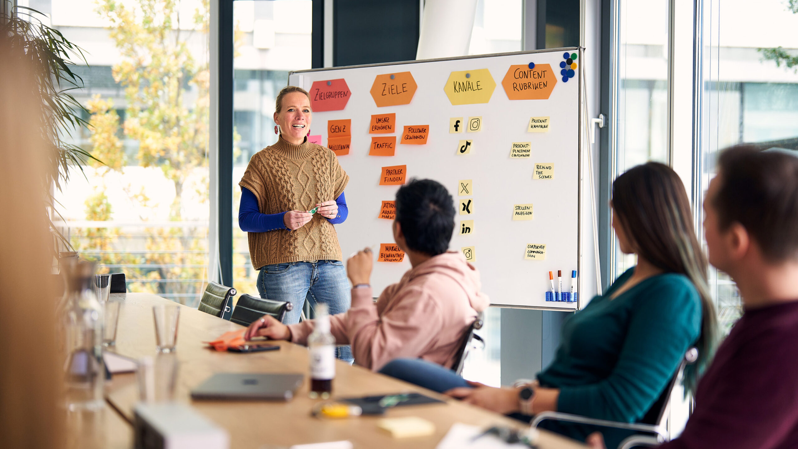 blonde Frau vor Whiteboard präsentiert in einem Konfi vor ihren Kolleg*innen