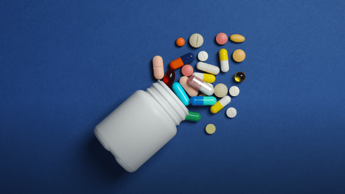 Eine weiße Dose liegt auf blauem Untergrund, bunte Pillen fallen heraus